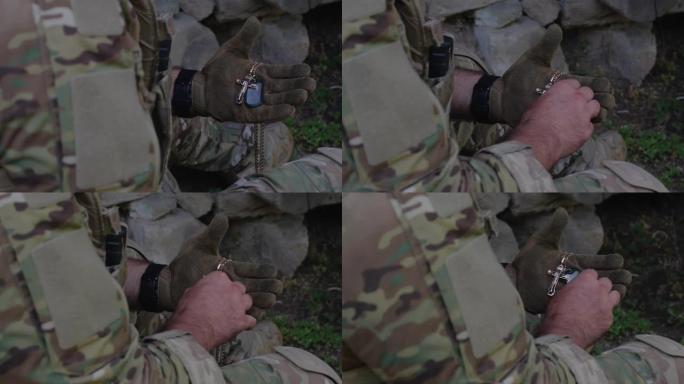 一名军人身穿迷彩服，戴着战术手套，手里拿着徽章和金属链上的十字架，在战斗前把它压在胸前，压在心上祈祷