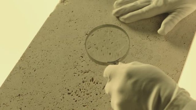 施工实验室检测天然钙华石的质量密度。实验室工作人员用放大镜探测石头。戴手套的手的特写视图。由ARRI