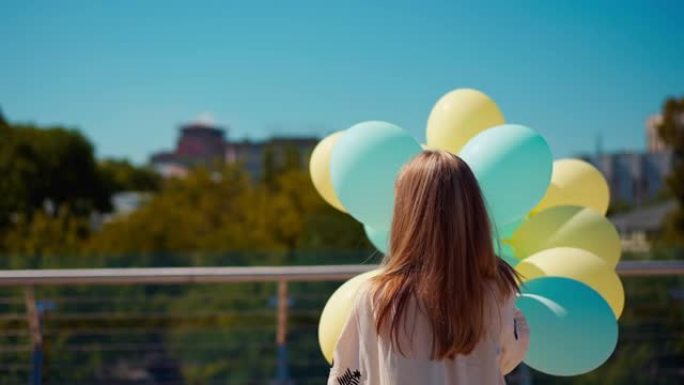 背面视图黑发少女穿着刺绣乌克兰衬衫，蓝色和黄色气球站在乌克兰基辅的玻璃桥上。青少年在阳光下的公园和城