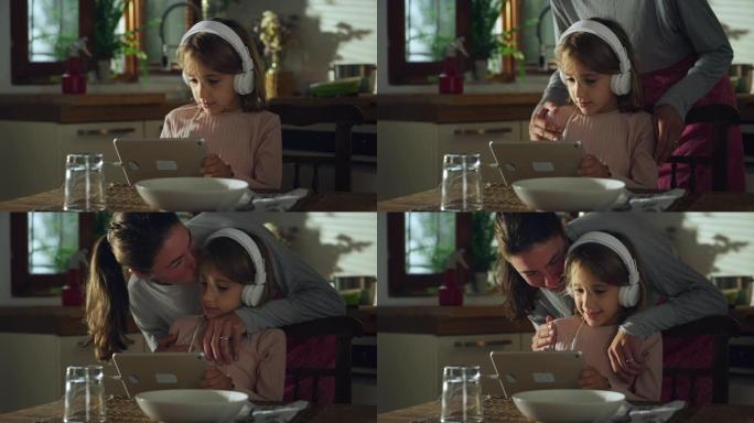 一个可爱的小女孩在厨房里使用数字平板电脑的肖像。当母亲在午餐时间来检查她时，玩教育游戏和使用技术的女