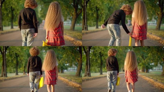 后视图礼貌的高加索男孩帮助女孩拿文件夹和朋友在户外散步。小绅士在公园小巷漫步，妹妹微笑着。童年和教育