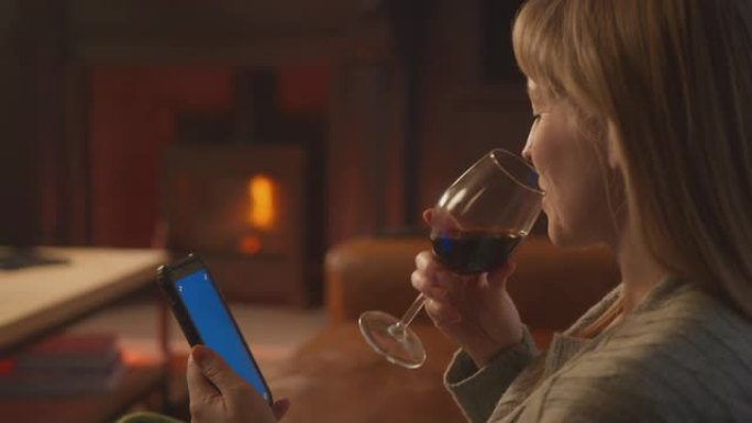 在家中的女人坐在沙发上带火用蓝屏手机拿着一杯酒
