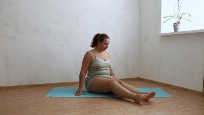 昏昏欲睡的超重女人坐在空荡荡的房间里的蓝色体操垫上，上下摇晃双腿，免费复制空间。家庭晨练，热身，运动