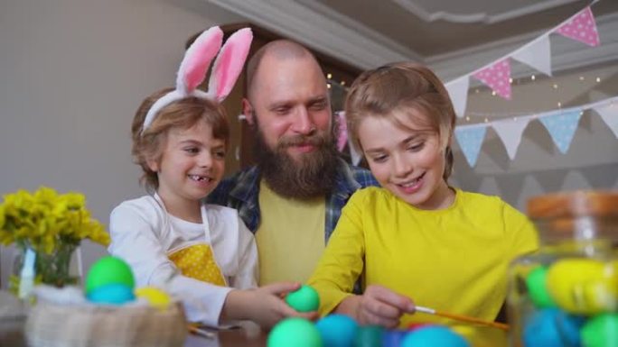 复活节家庭传统。父亲和两个白种人快乐的孩子，兔子耳朵在家里坐在一起的桌子上玩复活节彩蛋，用油漆过节。