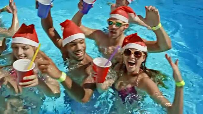 游泳池里的SLO MO朋友拿着他们的饮料，头上戴着圣诞老人的帽子向镜头挥手