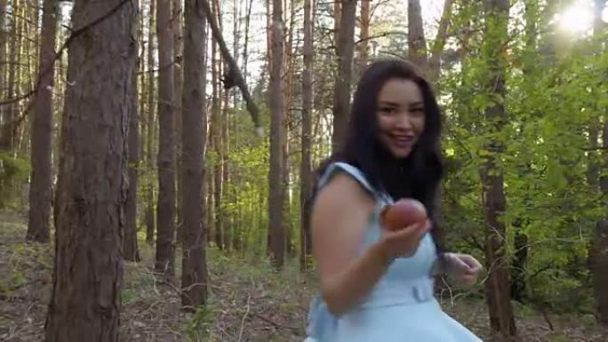 穿着蓝色连衣裙的迷人黑发女人手里拿着苹果走在森林里