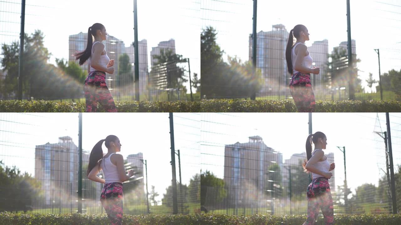 侧视图适合年轻的女运动员在阳光下慢动作地沿着网状栅栏奔跑。现场摄像机跟随苗条的白人运动女子在户外阳光