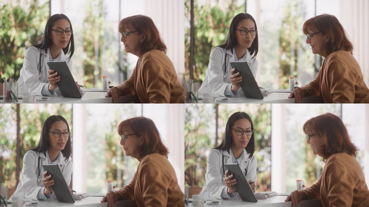 年轻的亚洲全科医生在健康诊所咨询期间向高级女性患者在平板电脑上显示心脏分析结果。医生坐在医院办公室的
