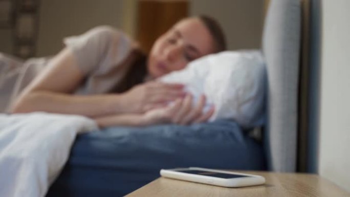 是时候醒来了。清晨的概念。躺在床上的一个快乐的女人伸出手来关闭智能手机屏幕上的闹钟。