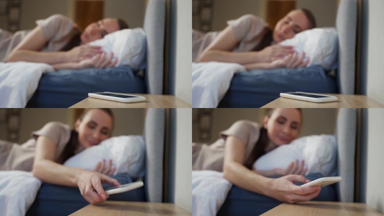 是时候醒来了。清晨的概念。躺在床上的一个快乐的女人伸出手来关闭智能手机屏幕上的闹钟。