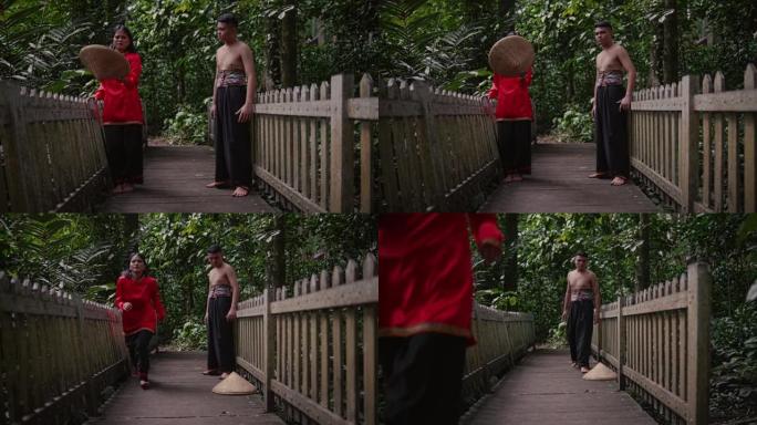 一位穿着红色衣服的妇女在竹桥中间向伴侣扔了一顶竹帽，表情愤怒
