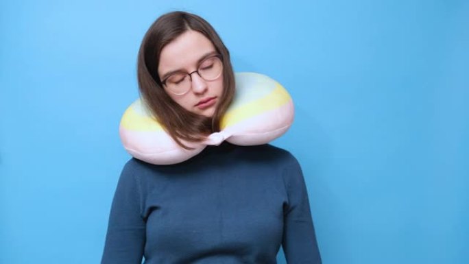 视频肖像: 一个戴着眼镜的学生女孩和一件穿着旅行枕头的毛衣，在蓝色背景上睡觉，4K。一个带着颈枕的年