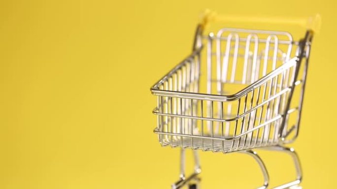 空的微型玩具超市手推车，旋转隔离在黄色背景上。现代社会消费主义的象征，缺乏金钱，商品。
