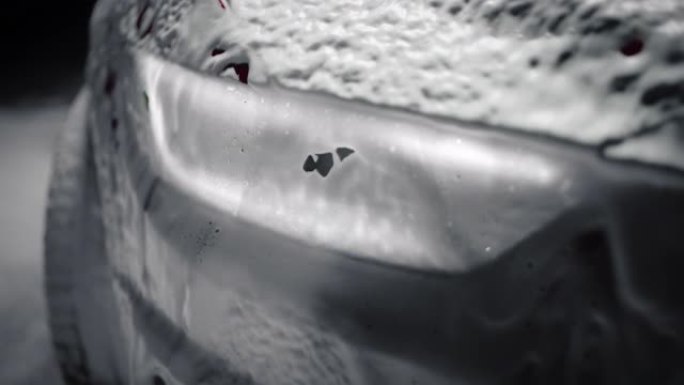 在黑暗的工作室环境中，用白色蓬松洗发水覆盖的现代汽车的特写镜头。在细节洗车过程中，表演跑车滴落在泡沫