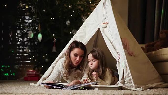 我的母亲和女儿穿着白色毛衣，与她躺在一个已安装房屋的帐篷里的夜晚读了一个童话故事。女孩笑，妈妈笑