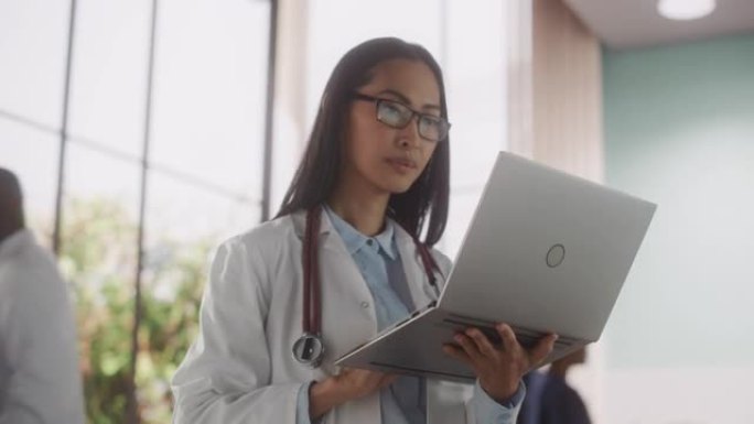 年轻的女医生穿着白大褂在医院散步和使用笔记本电脑。美丽的亚洲医疗保健专业人士在线治疗患者。动态手持镜