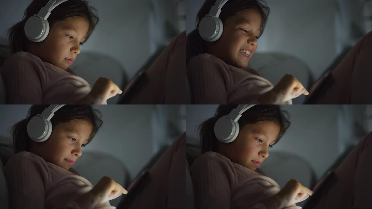 一个小女孩在睡觉前使用耳机和数字平板电脑玩游戏的特写镜头。女孩子晚上专注于屏幕，看视频，笑，玩得开心