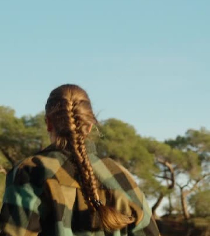 一位穿着格子衬衫的辫子的年轻女子在日落时分穿过森林。后视图-慢动作垂直视频社交媒体