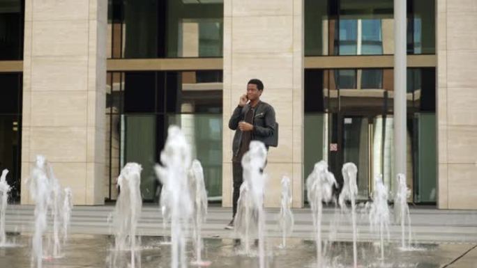 年轻的非洲男子站在喷泉后面的大楼附近，用手机、手里拿着咖啡杯和胳膊下的笔记本电脑交谈