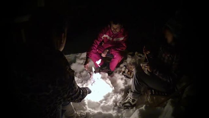 一群人晚上在雪地上燃烧篝火，在棍子上炸香肠