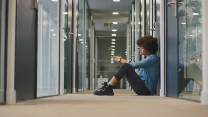 紧张的年轻女商人坐在带电话的现代办公室走廊的地板上