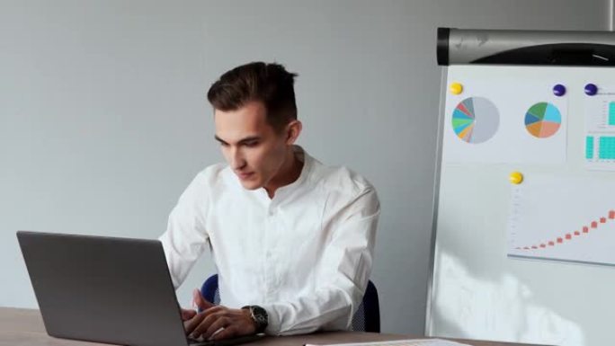 一位欧洲男性的肖像坐在办公室的笔记本电脑上，白色衬衫在图表和桌子的背景上