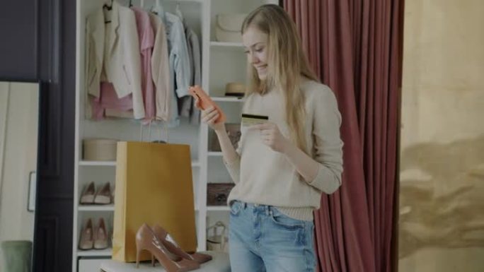 快乐的女人使用智能手机和信用卡站在衣柜和纸袋旁边在线购物
