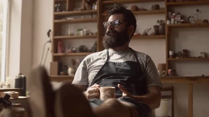 戴着眼镜的大胡子时尚男陶工坐在他的陶器车间里喝自己生产的一杯咖啡，喝咖啡休息，工作时休息