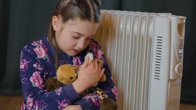 客厅冷冻小女孩在电暖器散热器和拥抱毛绒玩具旁边取暖