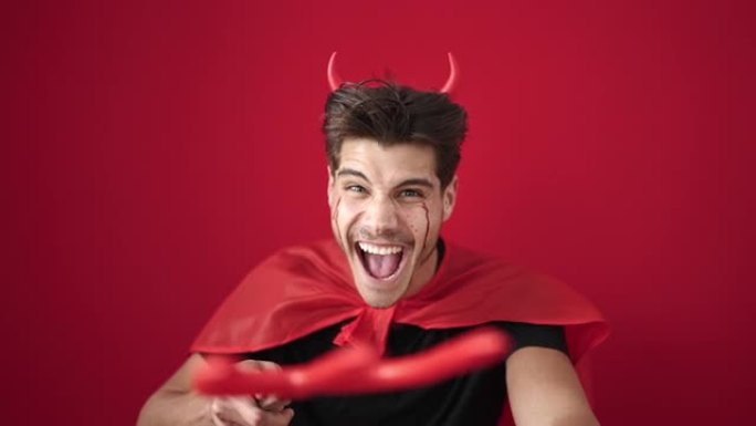 年轻的西班牙裔男子穿着恶魔服装，三叉戟在孤立的红色背景上袭击