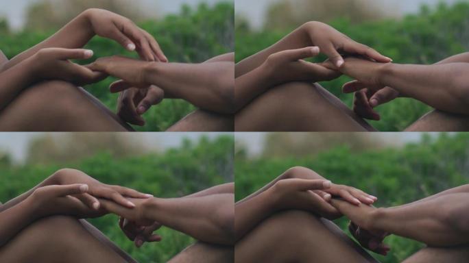 特写手。非洲族裔浪漫情侣在旷野约会。坐在草地上牵着手