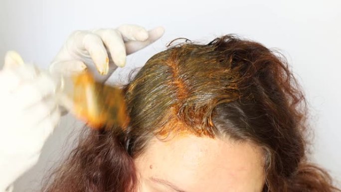 剪下的美发师戴着白手套的照片，在客户的深棕色长发的根部涂上橙色染发剂。