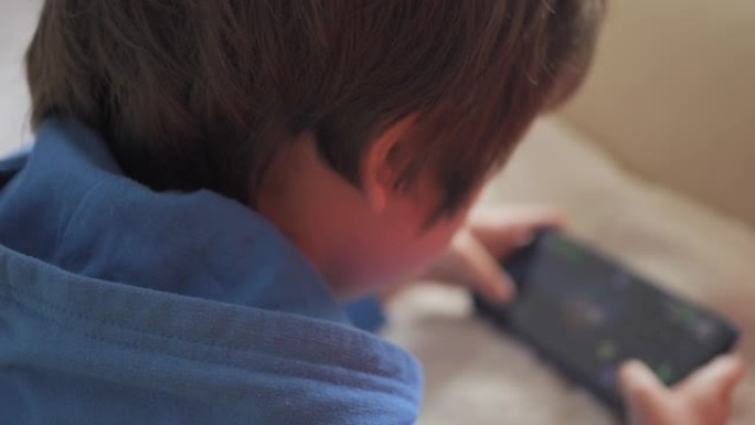 孩子躺在沙发上在家打电话玩游戏。男孩在手机上玩电子游戏。学龄前儿童在沙发上玩电子游戏智能手机。孩子使