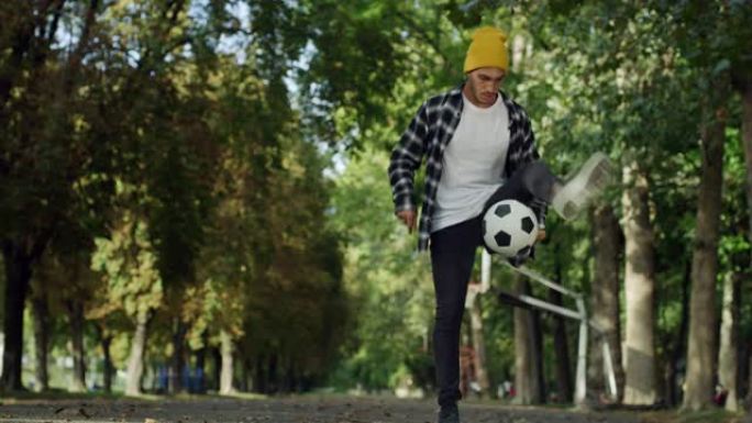 才华横溢的年轻足球运动员熟练地玩弄球，在阳光明媚的日子里在公共场合表演。时髦的男人在公园里炫耀自己的