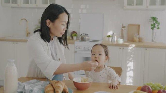 慈爱的母亲在公寓的厨房里用果泥喂养小女儿照顾孩子