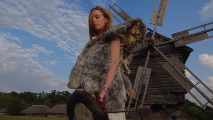 一个金发女孩穿着用动物皮制成的异教毛皮服装，拿着一个皮手鼓站在一个古老的木制风车和乡村风景的背景下。