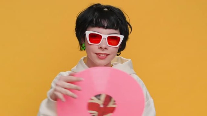 音乐唱片乙烯基复古粉色盘子快乐少女跳舞唱歌旋转握在手中，戴着墨镜把它带到相机前，在黄色背景上看起来直