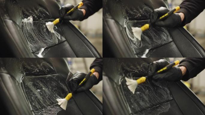 汽车细节和洗车概念。男人，洗车工人，穿着黑色衣服和防护橡胶手套，清洁汽车前排座椅，将泡沫喷在软刷上