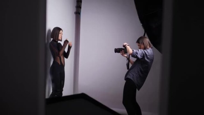 穿着黑色紧身西装和皮靴的性感年轻时尚女性模特在摄影棚里与摄影师在白色背景上合影。后台