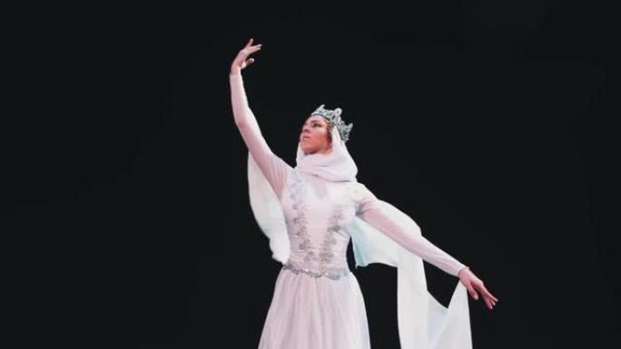 穿着漂亮的白色西装和皇冠的芭蕾舞演员在黑色背景的剧院舞台上表演美丽的芭蕾舞元素。风吹着她的衣服。举起