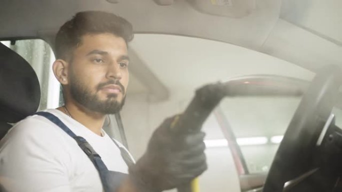 用蒸汽清洁器清洁汽车沙龙。专业男性自信工人使用热蒸汽清洁器清洁汽车内饰、方向盘和控制面板的侧视图。清