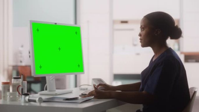一名黑人女性医疗保健专业人员的肖像在医院办公室的台式电脑上工作，绿屏模拟显示。诊所护士长正在在线指定