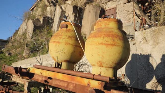 山坡石前球形的旧煤气罐。两个旧的圆形黄色气瓶站在山坡上建在岩石中的石头住宅前的铁基座上