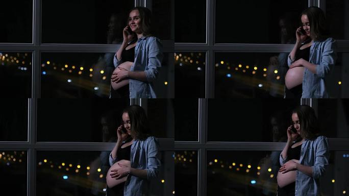 一名孕妇在夜里用手机说话双手摸着肚子站在窗前