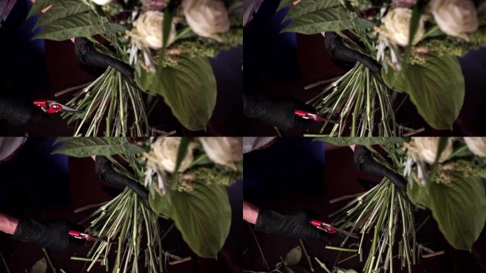 花匠戴着黑色防护手套的手的特写镜头用红色剪刀剪掉了花束。花店收集一束白玫瑰