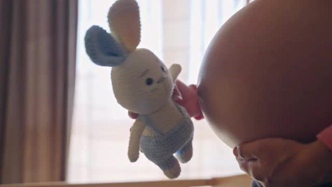 未来妈妈手中的针织兔子玩具在窗户背景下亲吻怀孕的肚子，美丽的孕妇期待她的孩子，产前的幸福