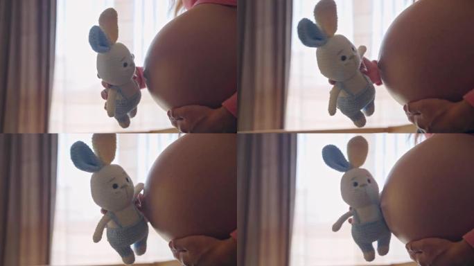 未来妈妈手中的针织兔子玩具在窗户背景下亲吻怀孕的肚子，美丽的孕妇期待她的孩子，产前的幸福