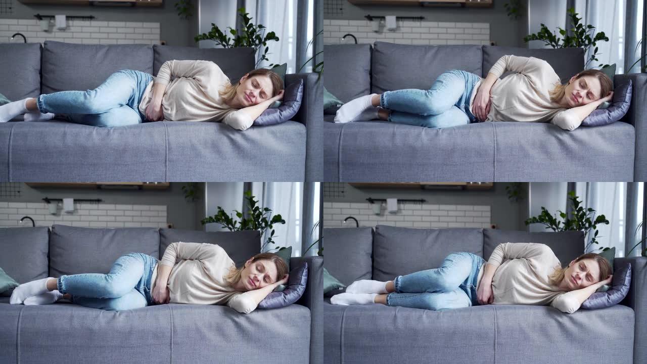 生病的年轻金发女子患有胃痛躺在沙发上抱着肚子在家里的客厅感觉胃炎或月经疼痛