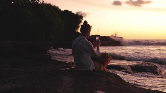 坐在夏日海滩上的女人使用手机并拍摄海洋自然的照片。夏季度假的成年女孩在户外看智能手机的概念。黎明时海