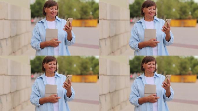 微笑的中年妇女50-60岁使用智能手机，并在温暖的日子在户外拿着教科书。成年学生在校园学院之间有一段